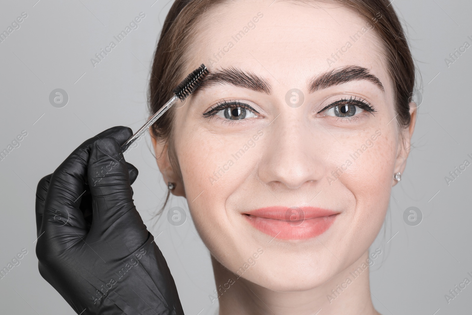 Photo of Brow lamination. Cosmetologist combing woman's eyebrows with brush against grey background, closeup