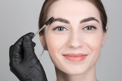 Photo of Brow lamination. Cosmetologist combing woman's eyebrows with brush against grey background, closeup