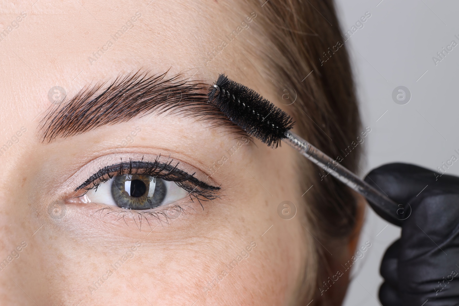 Photo of Brow lamination. Cosmetologist combing woman's eyebrows with brush against grey background, closeup