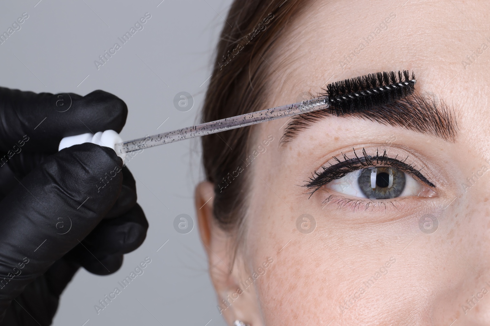 Photo of Brow lamination. Cosmetologist combing woman's eyebrows with brush against grey background, closeup