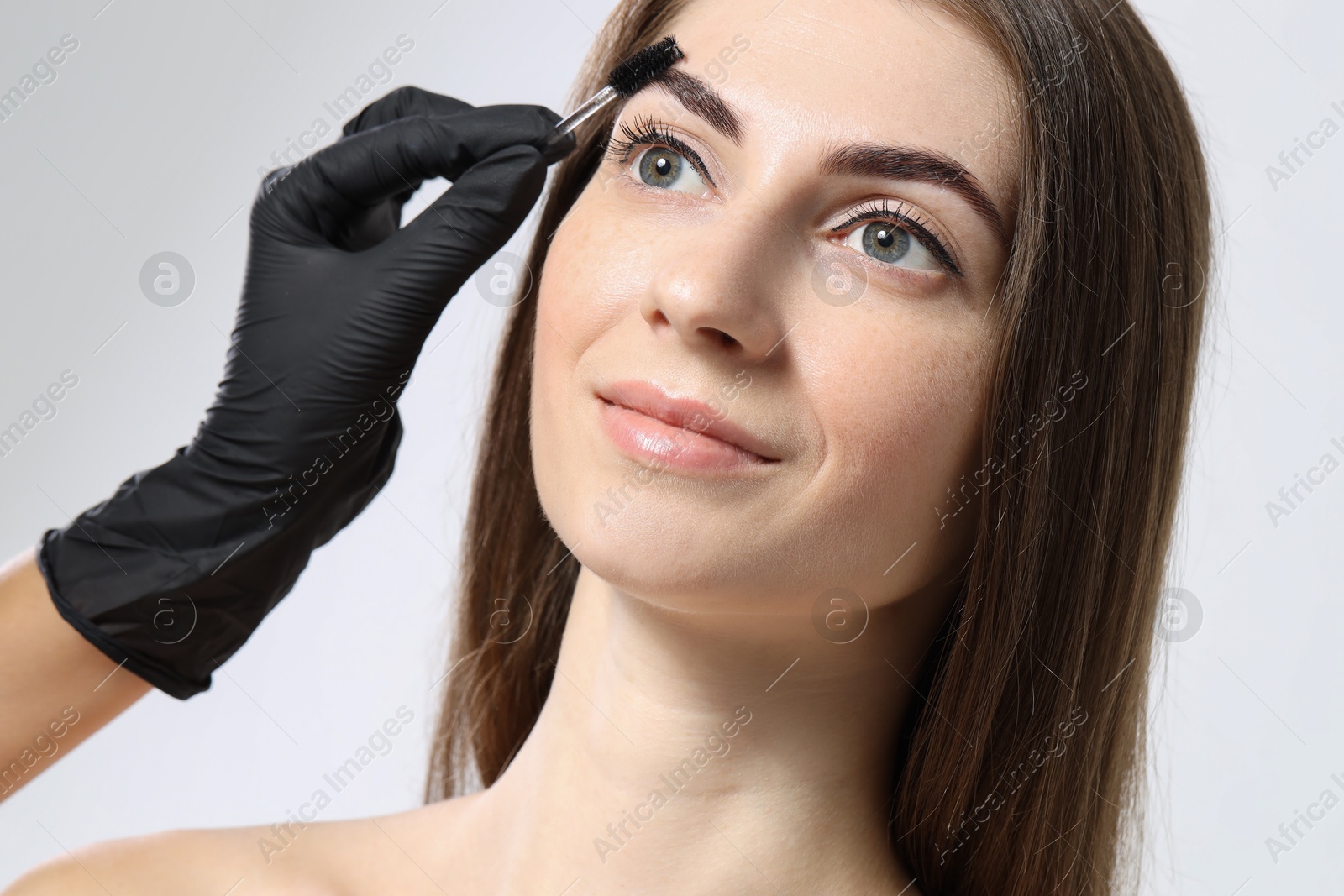 Photo of Beautician brushing client's eyebrows after henna dyeing procedure on light background, closeup