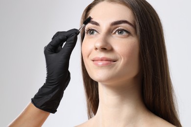 Photo of Beautician brushing client's eyebrows after henna dyeing procedure on light background, closeup