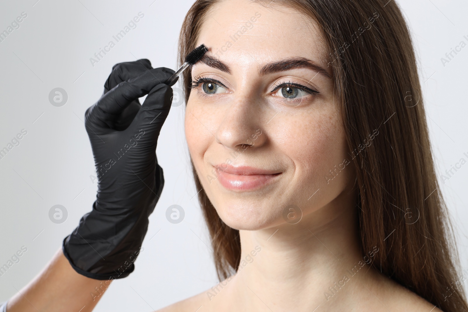 Photo of Beautician brushing client's eyebrows after henna dyeing procedure on light background, closeup