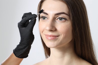 Photo of Beautician brushing client's eyebrows after henna dyeing procedure on light background, closeup