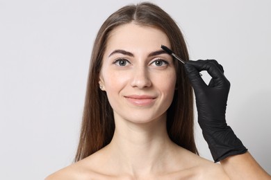 Photo of Beautician brushing client's eyebrows after henna dyeing procedure on light background, closeup