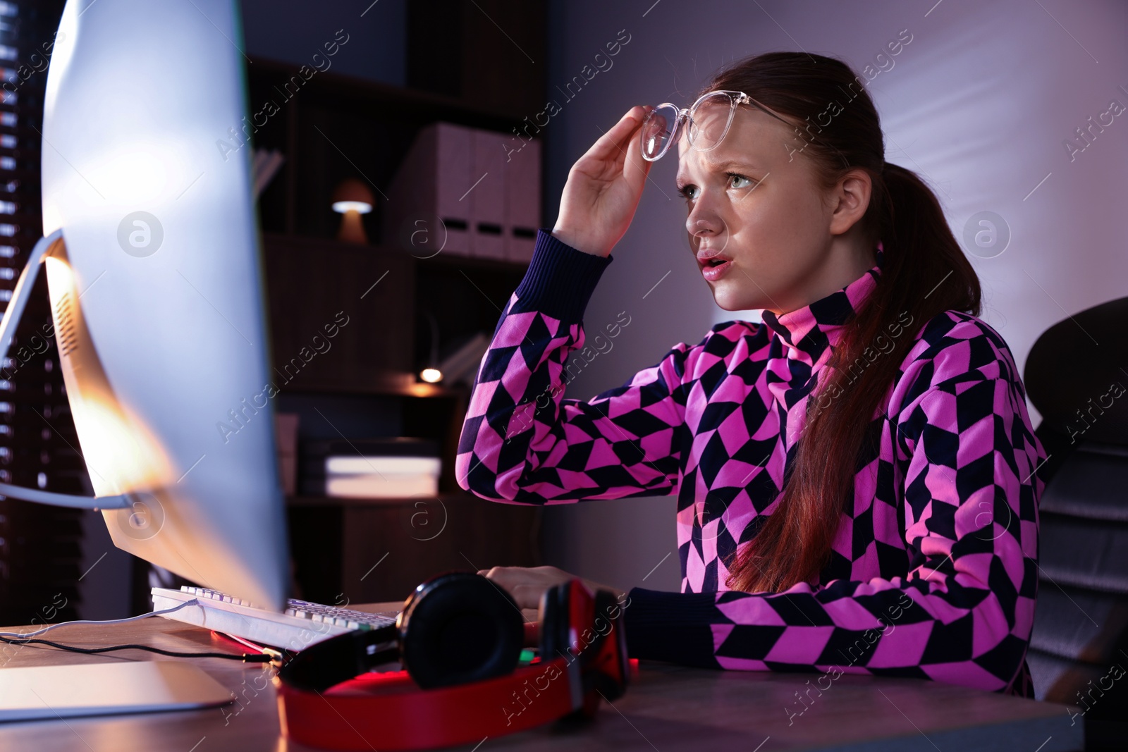 Photo of Girl looking at monitor at table with keyboard and headphones indoors