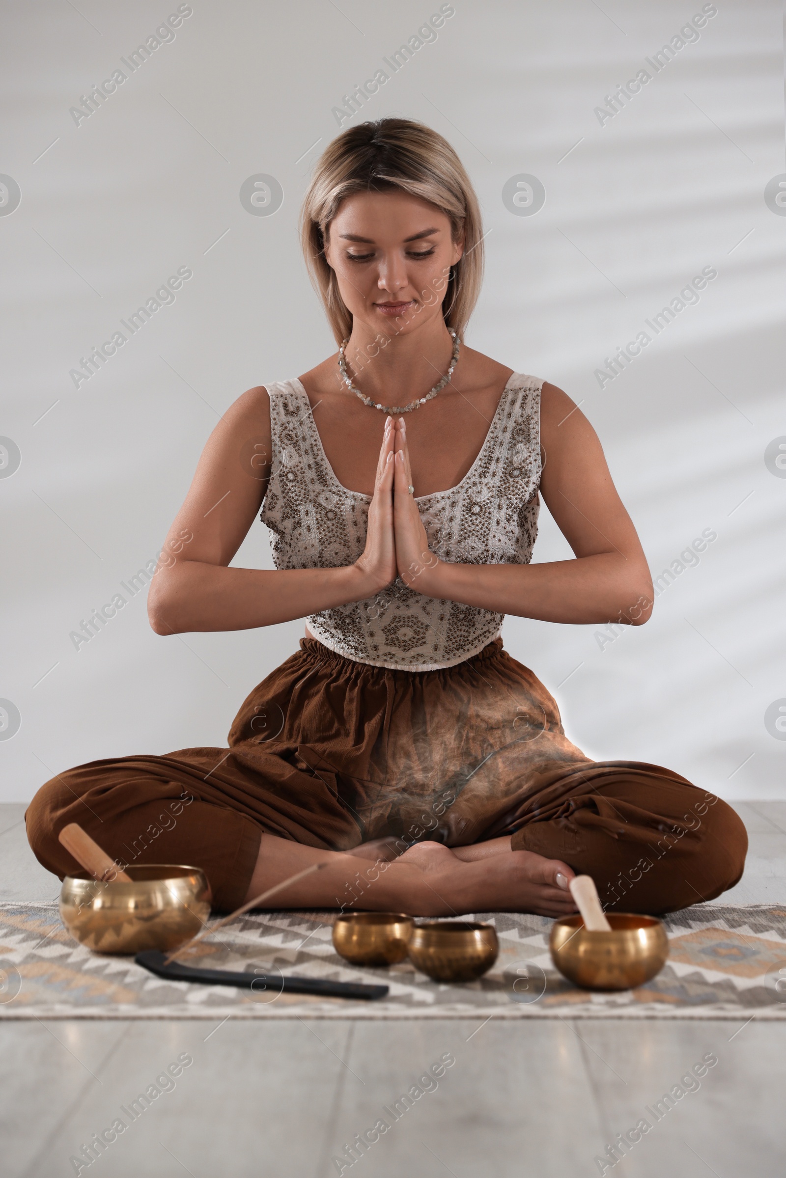 Photo of Beautiful young woman with smoldering incense stick and tibetan singing bowls meditating on floor indoors