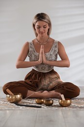 Beautiful young woman with smoldering incense stick and tibetan singing bowls meditating on floor indoors