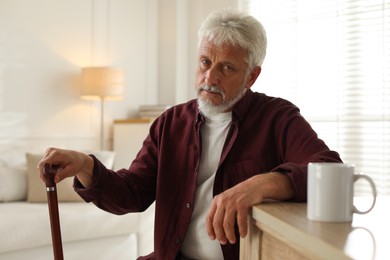 Photo of Lonely senior man with walking cane sitting at table in room