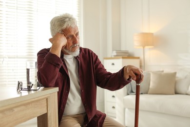 Photo of Lonely senior man with walking cane sitting at table in room