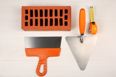 Photo of Red brick, putty knives and measuring tape on light wooden table, flat lay