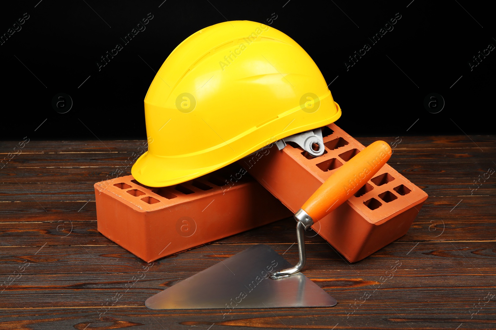 Photo of Red bricks, putty knife and yellow hard hat on wooden table. Building material