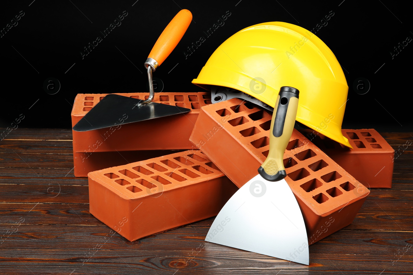 Photo of Red bricks, putty knives and yellow hard hat on wooden table. Building material