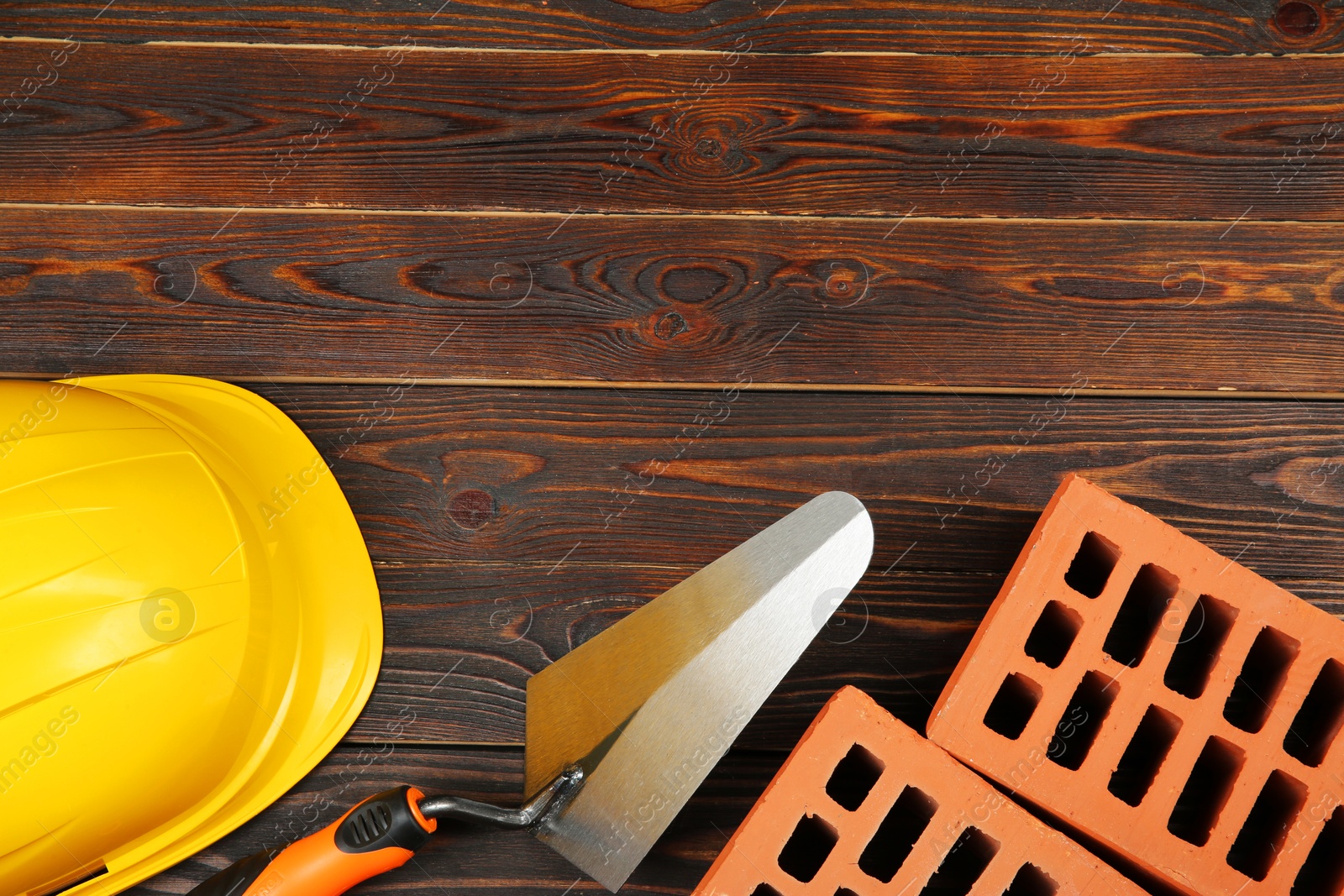 Photo of Red bricks, putty knife and yellow hard hat on wooden table, flat lay with space for text. Building material