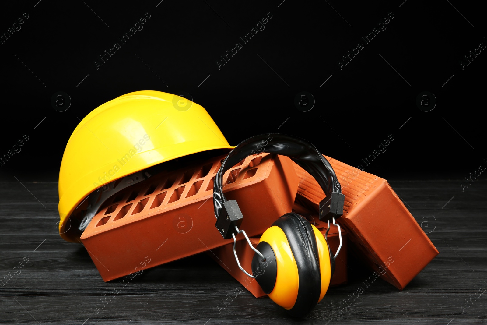 Photo of Red bricks, yellow hard hat and earmuffs on dark wooden table. Building material and safety equipment