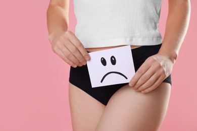 STD (sexually transmitted disease). Woman holding sticky note with drawn sad face on pink background, closeup