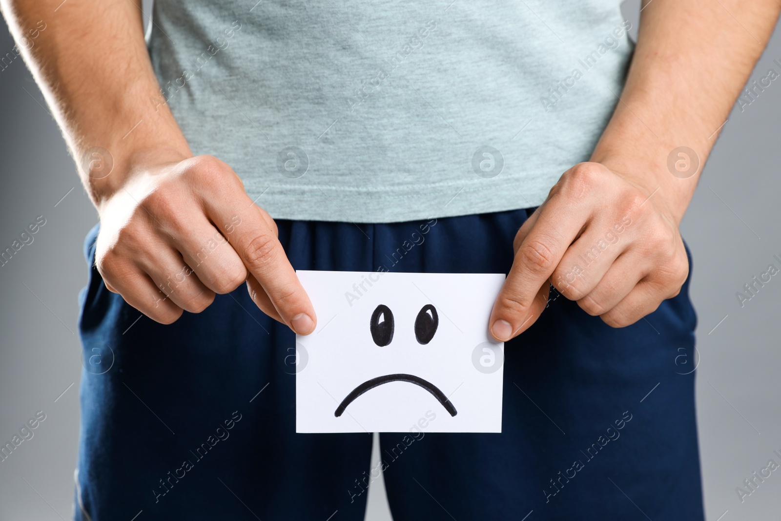 Photo of STD (sexually transmitted diseases). Man holding sticky note with drawn sad face on grey background, closeup