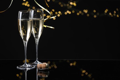 Photo of Champagne in glasses and party streamers on mirror surface against black background with blurred lights, space for text