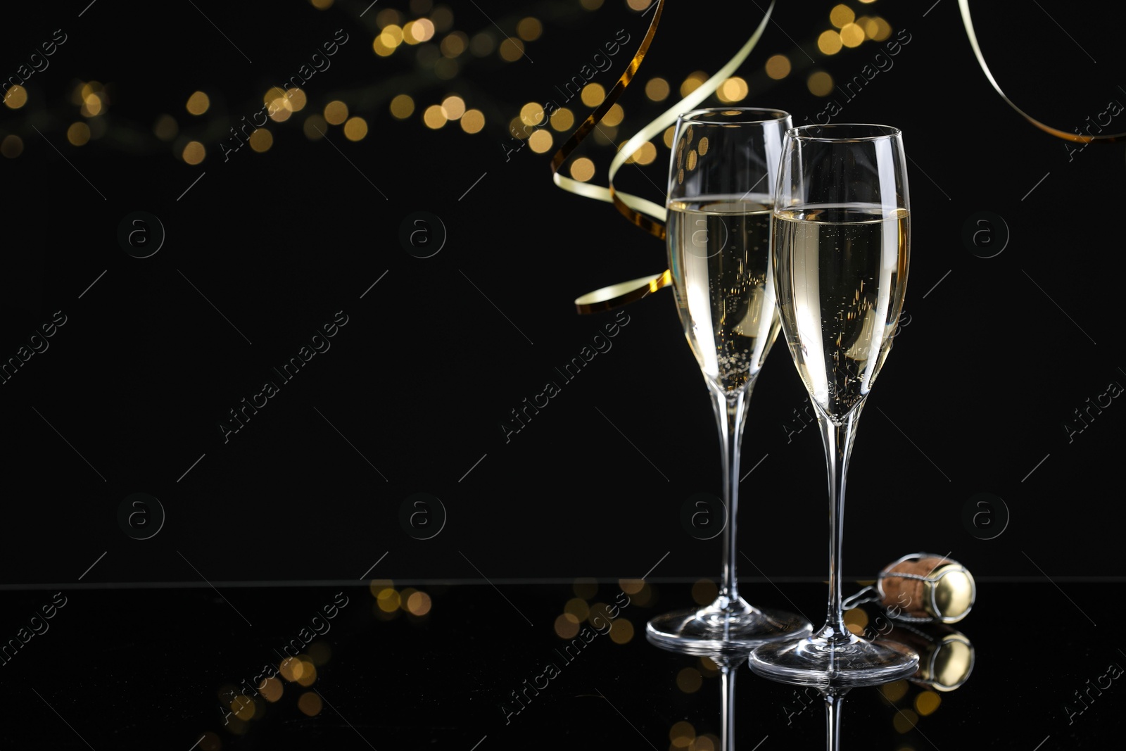 Photo of Champagne in glasses and party streamers on mirror surface against black background with blurred lights, space for text