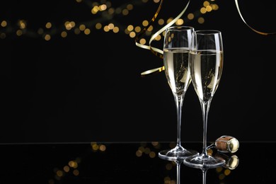 Photo of Champagne in glasses and party streamers on mirror surface against black background with blurred lights, space for text