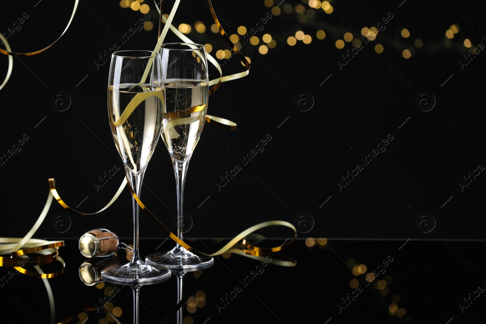 Photo of Champagne in glasses and party streamers on mirror surface against black background with blurred lights, space for text