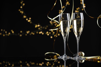 Photo of Champagne in glasses and party streamers on mirror surface against black background with blurred lights, space for text