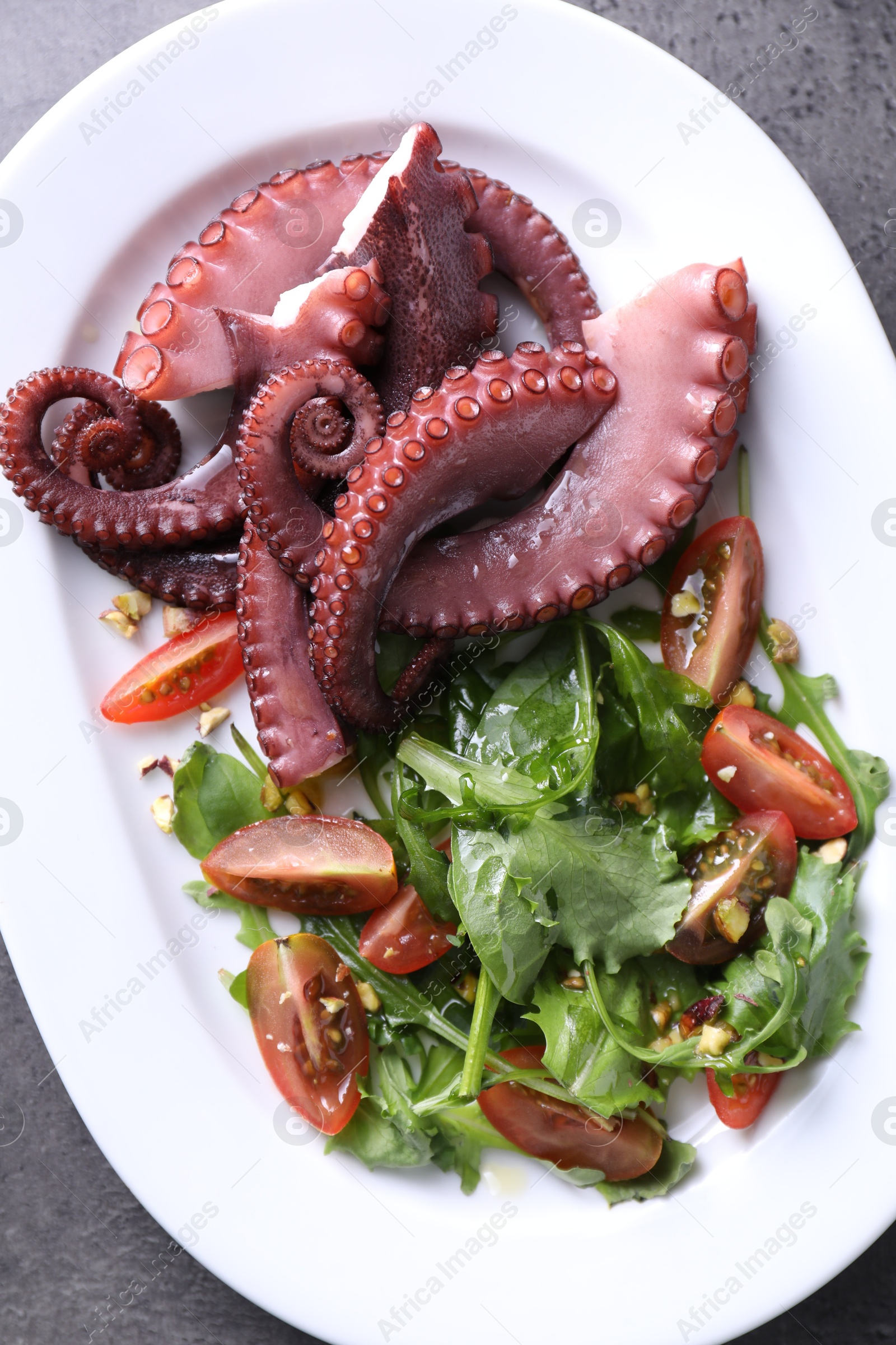 Photo of Plate with tasty boiled octopus tentacles and salad on grey textured table, top view