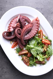 Photo of Plate with tasty boiled octopus tentacles and salad on grey textured table, top view