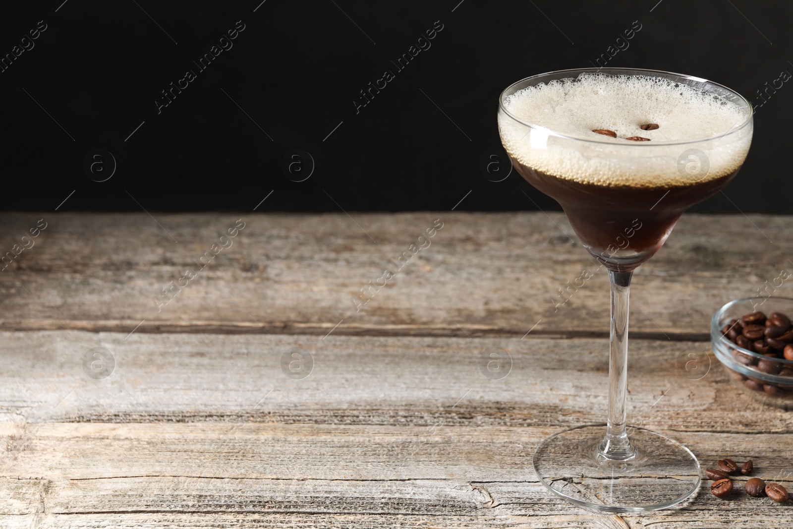 Photo of Glass of delicious espresso martini and coffee beans on wooden table against black background, closeup