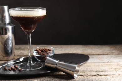 Photo of Glass of delicious espresso martini, jigger and coffee beans on wooden table against black background, closeup. Space for text