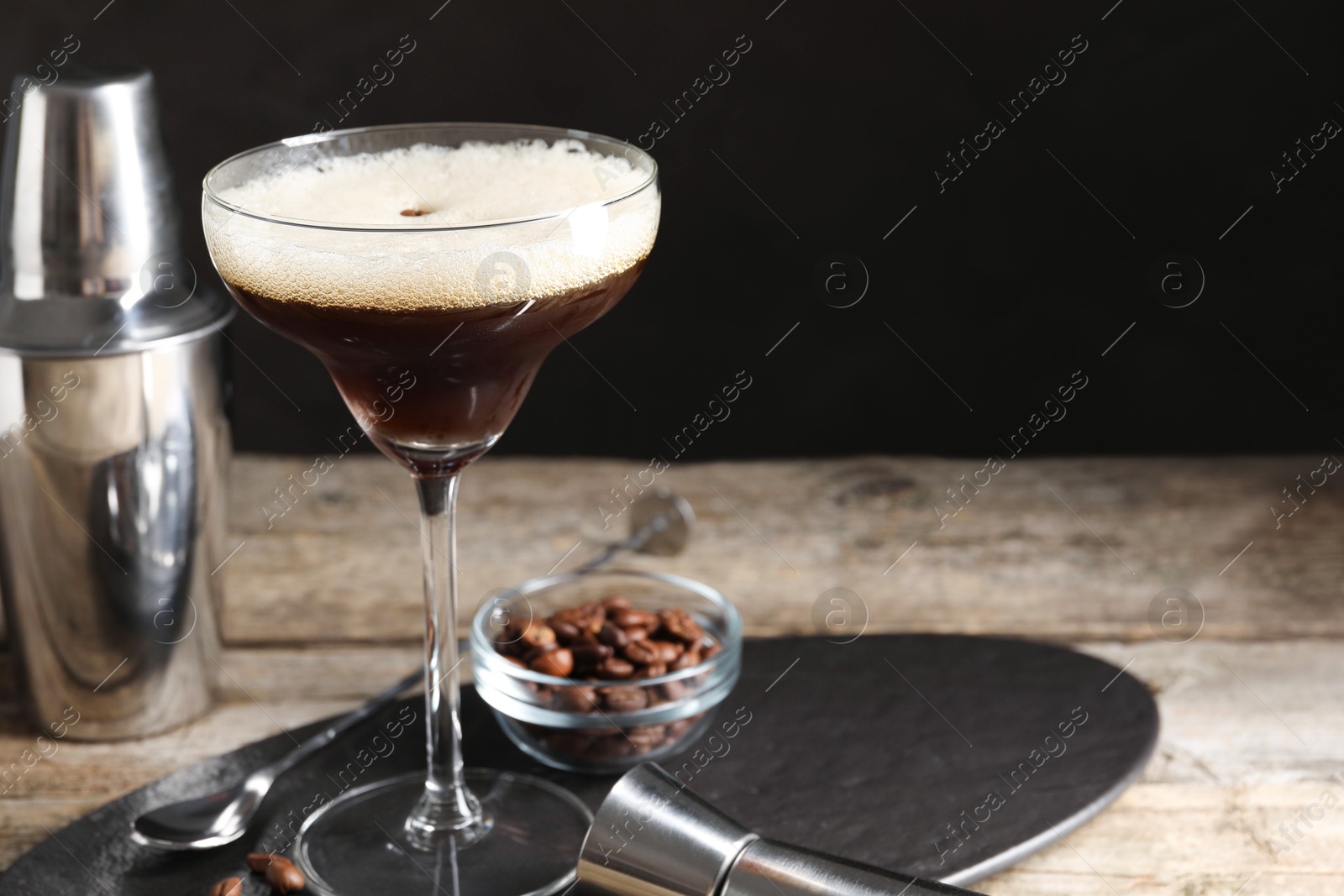 Photo of Glass of delicious espresso martini, jigger and coffee beans on wooden table against black background, closeup. Space for text