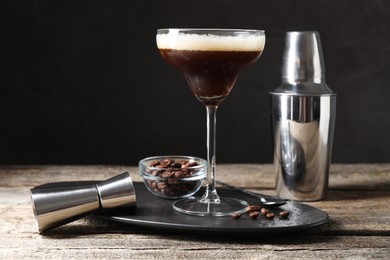 Glass of delicious espresso martini, jigger, shaker and coffee beans on wooden table against black background, closeup