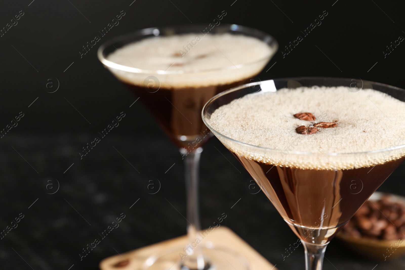 Photo of Delicious espresso martini with coffee beans in glasses on blurred black background, closeup