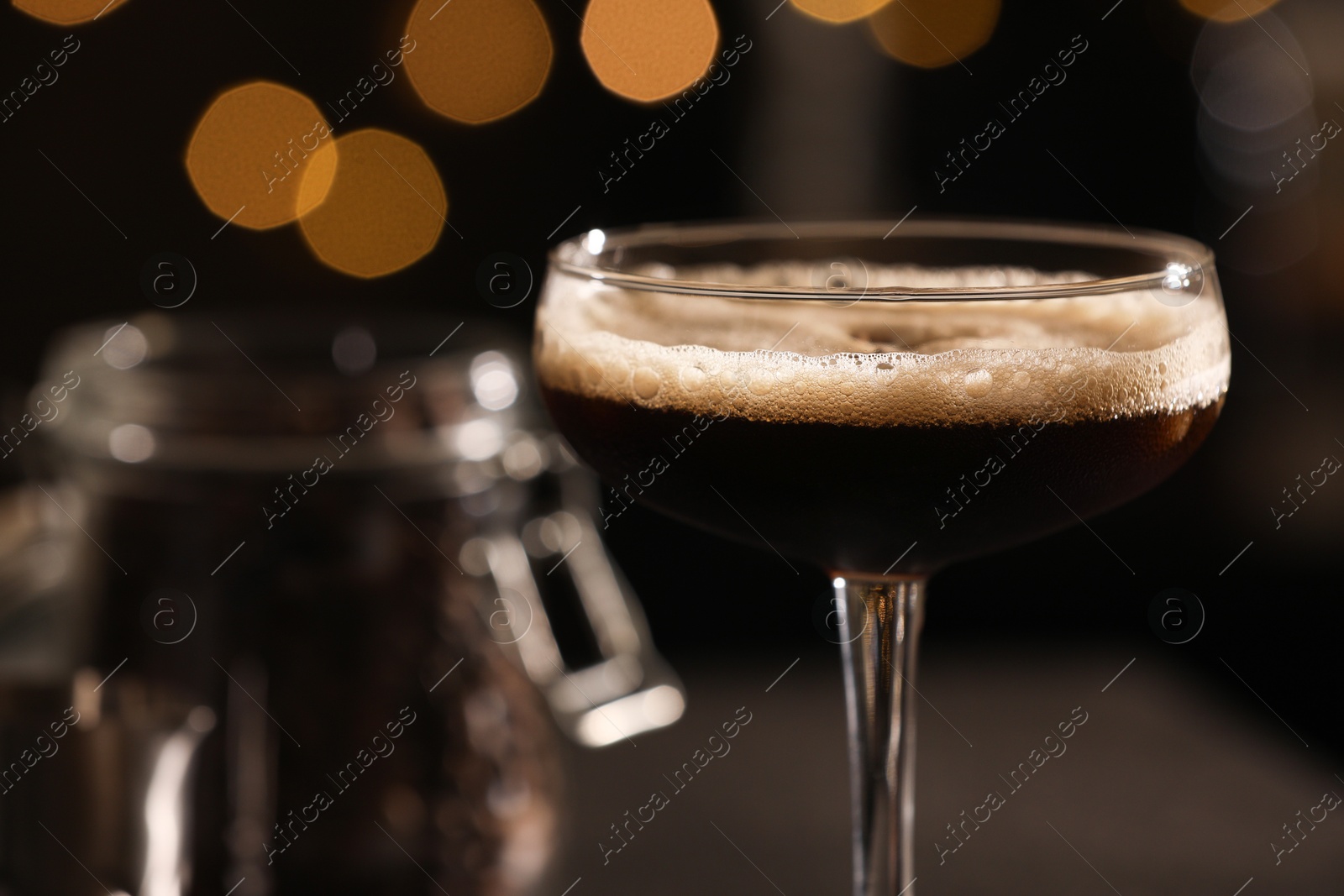 Photo of Delicious espresso martini in glass against blurred lights, closeup. Space for text