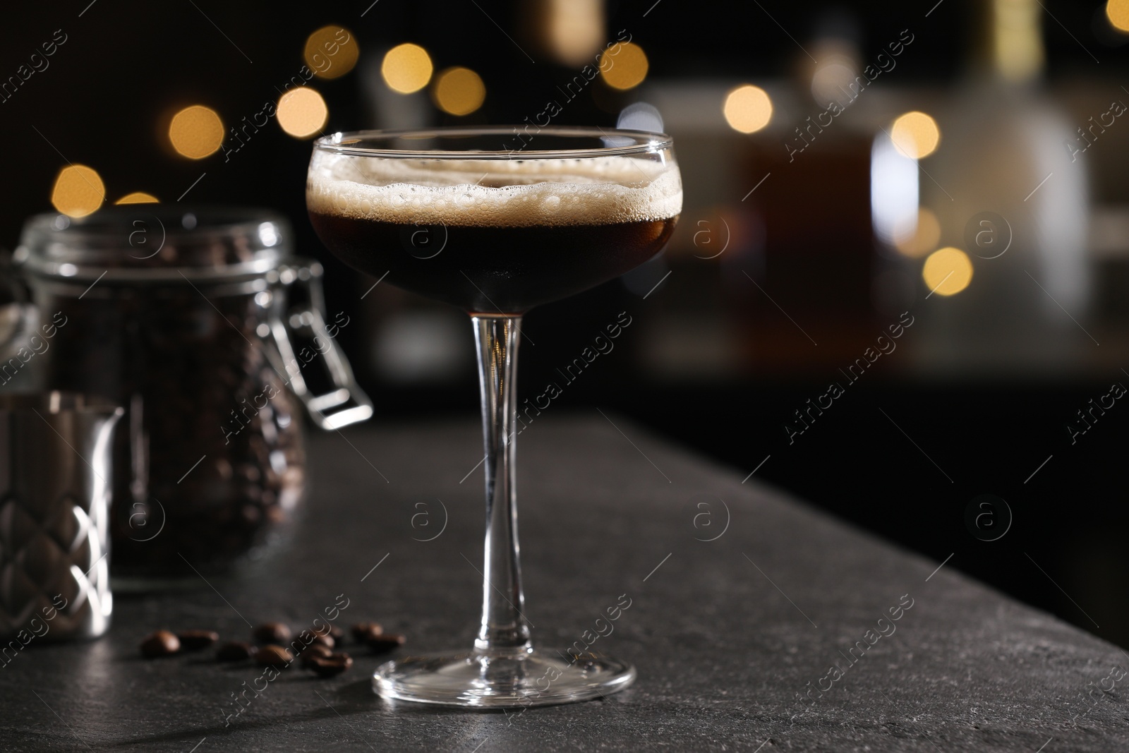 Photo of Glass with delicious espresso martini and coffee beans on dark table against blurred lights, closeup