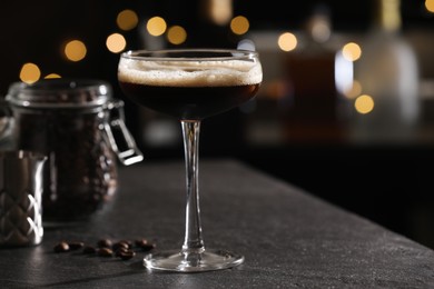 Photo of Glass with delicious espresso martini and coffee beans on dark table against blurred lights, closeup