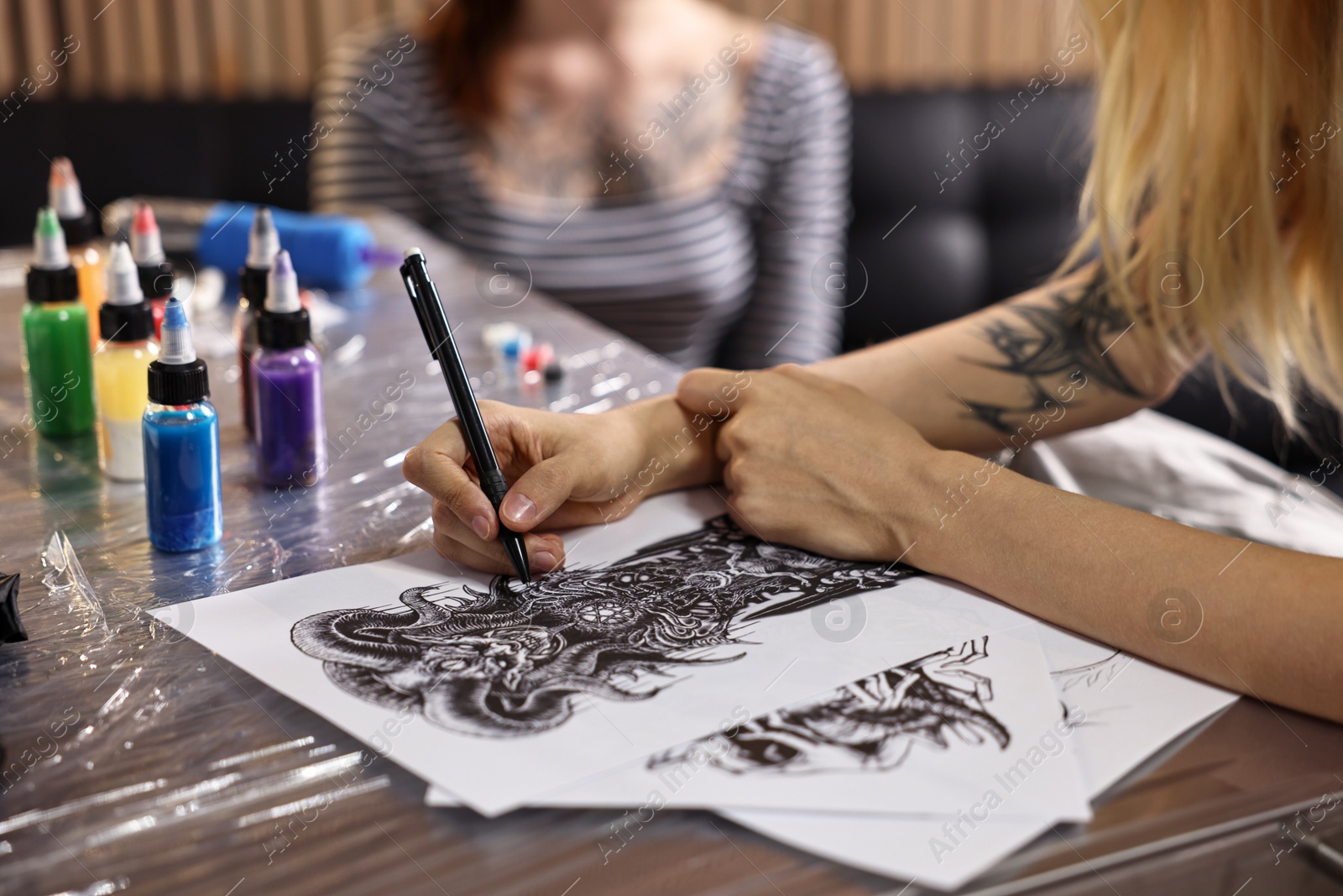 Photo of Tattoo artist drawing sketch at table with supplies near client in salon, closeup