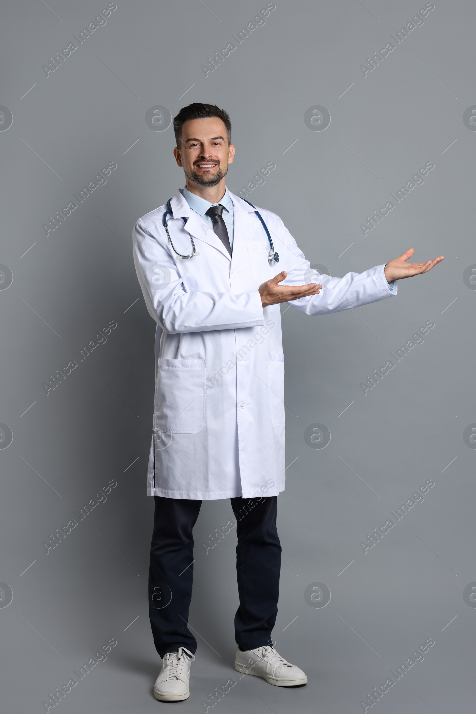 Photo of Smiling doctor with stethoscope pointing at something on grey background