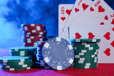 Photo of Poker chips and playing cards on pink table in neon lights, closeup