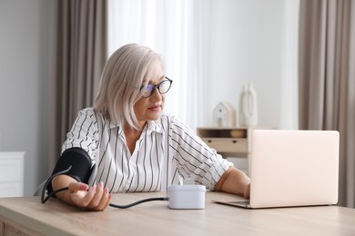 Photo of Woman measuring blood pressure while using laptop at wooden table indoors