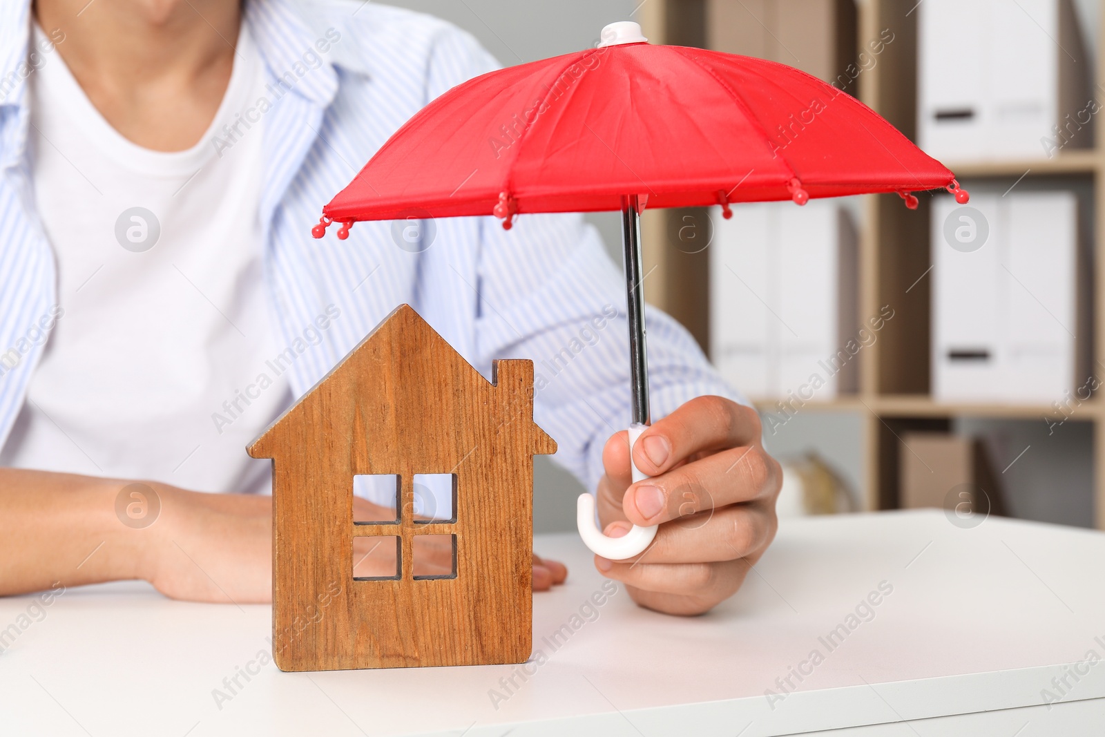 Photo of Real estate insurance. Man holding small umbrella above wooden house figure at light table, closeup