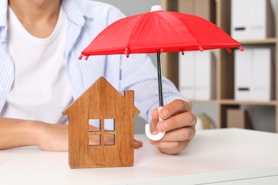 Photo of Real estate insurance. Man holding small umbrella above wooden house figure at light table, closeup