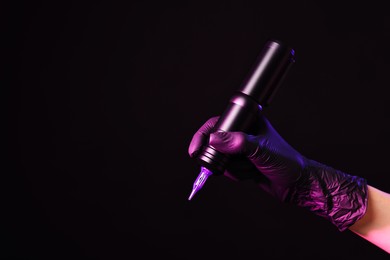 Photo of Professional artist holding tattoo machine on black background in color lights, closeup. Space for text