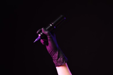 Photo of Professional artist holding tattoo machine on black background in color lights, closeup