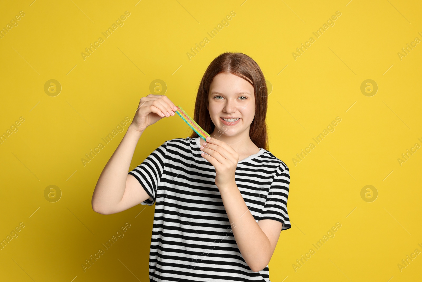 Photo of Happy teenage girl with tasty rainbow sour belt on yellow background