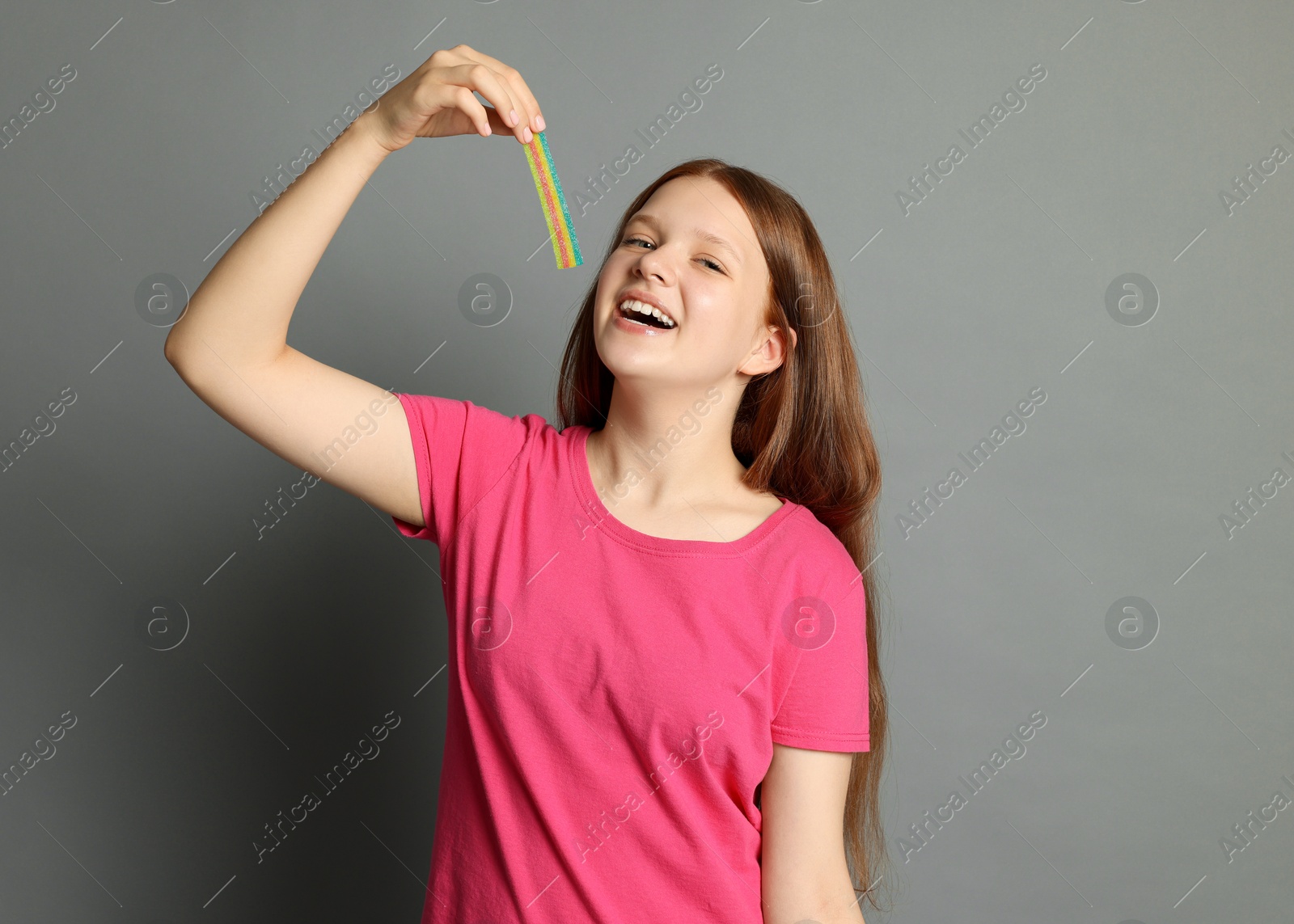 Photo of Happy teenage girl eating tasty rainbow sour belt on grey background