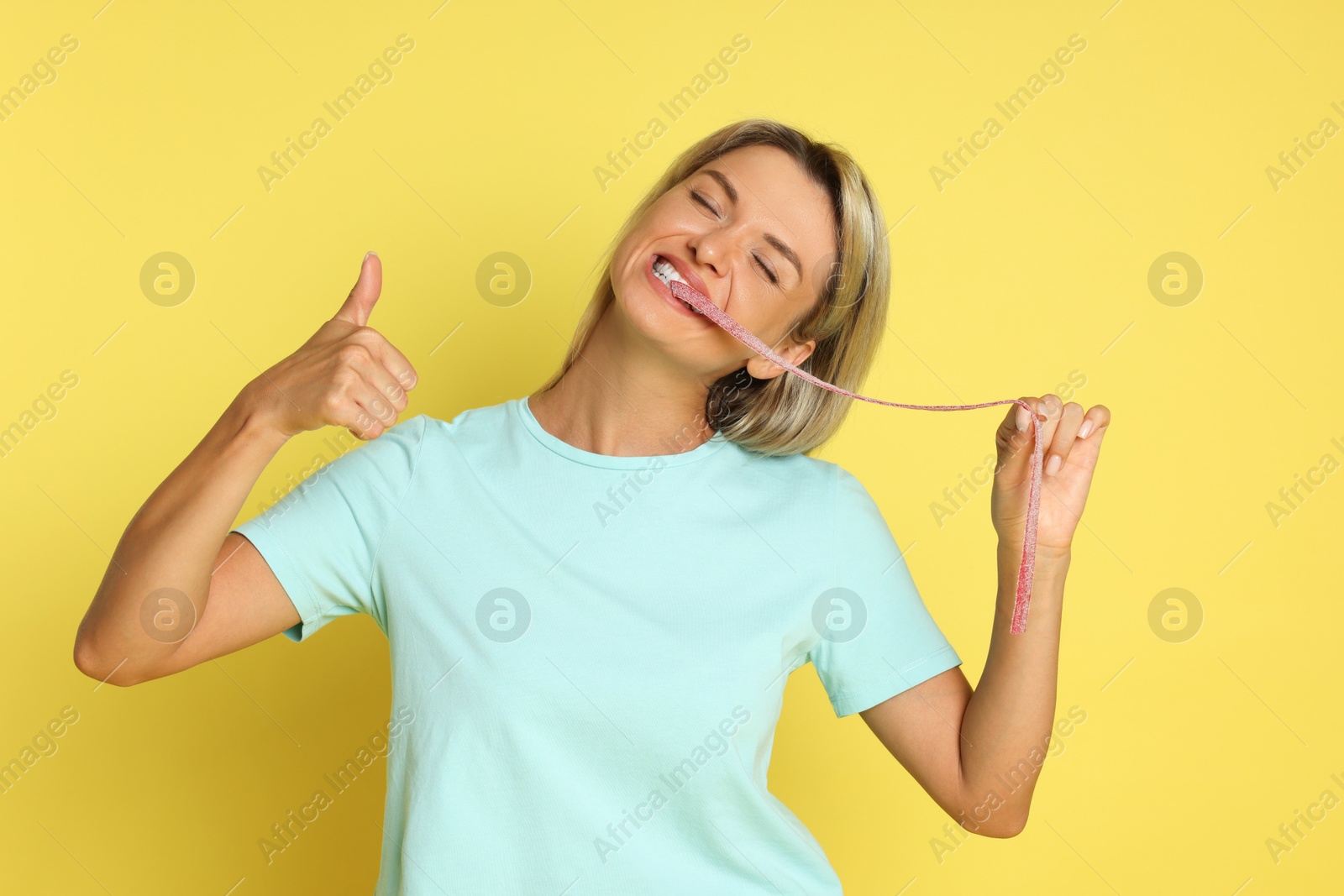 Photo of Young woman eating tasty gummy candy while showing thumbs up on yellow background