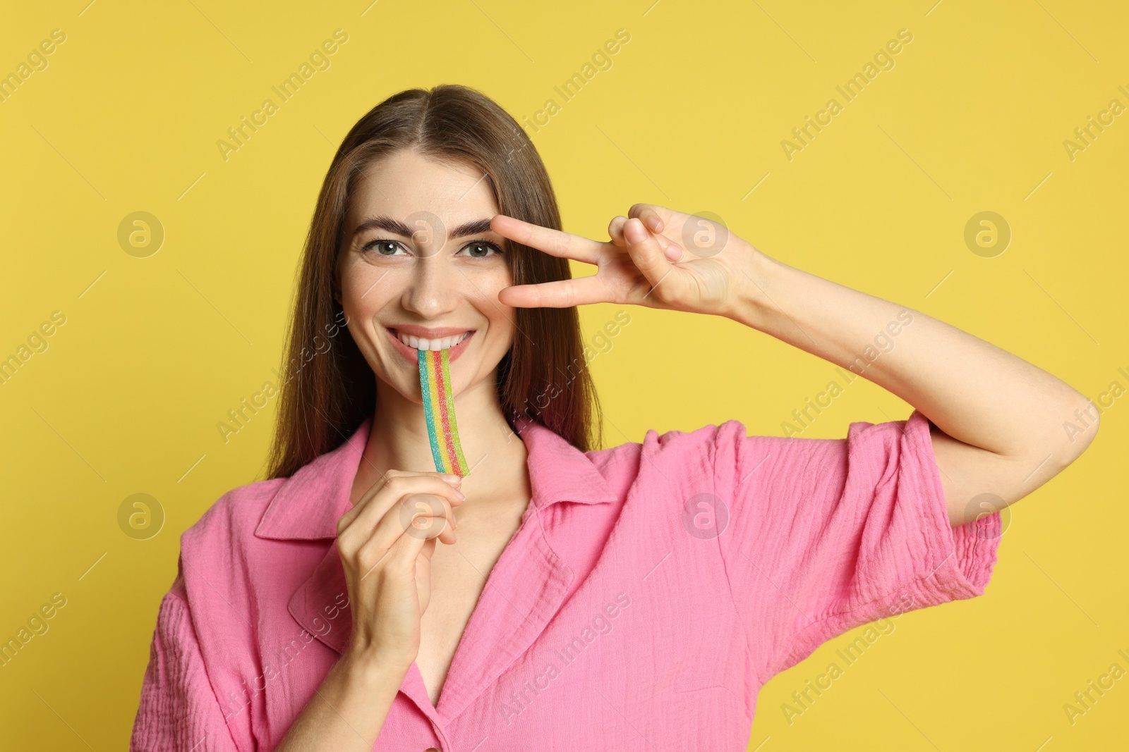 Photo of Young woman eating tasty rainbow sour belt while showing v-sign on yellow background