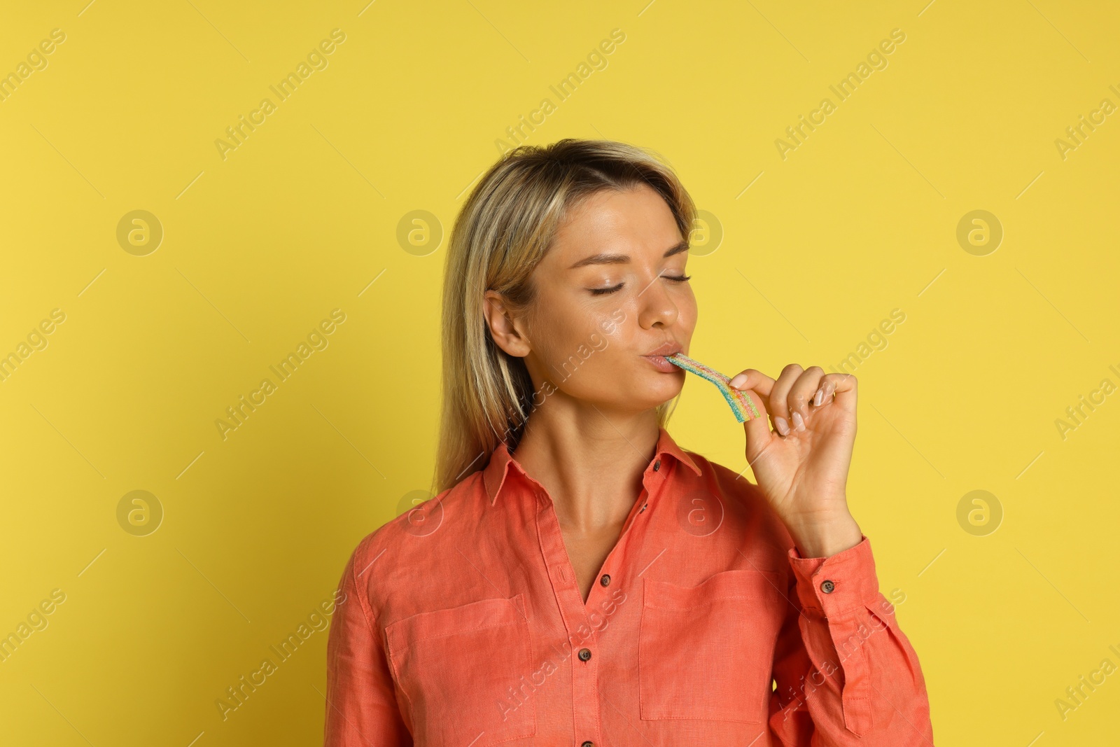 Photo of Young woman eating tasty rainbow sour belt on yellow background