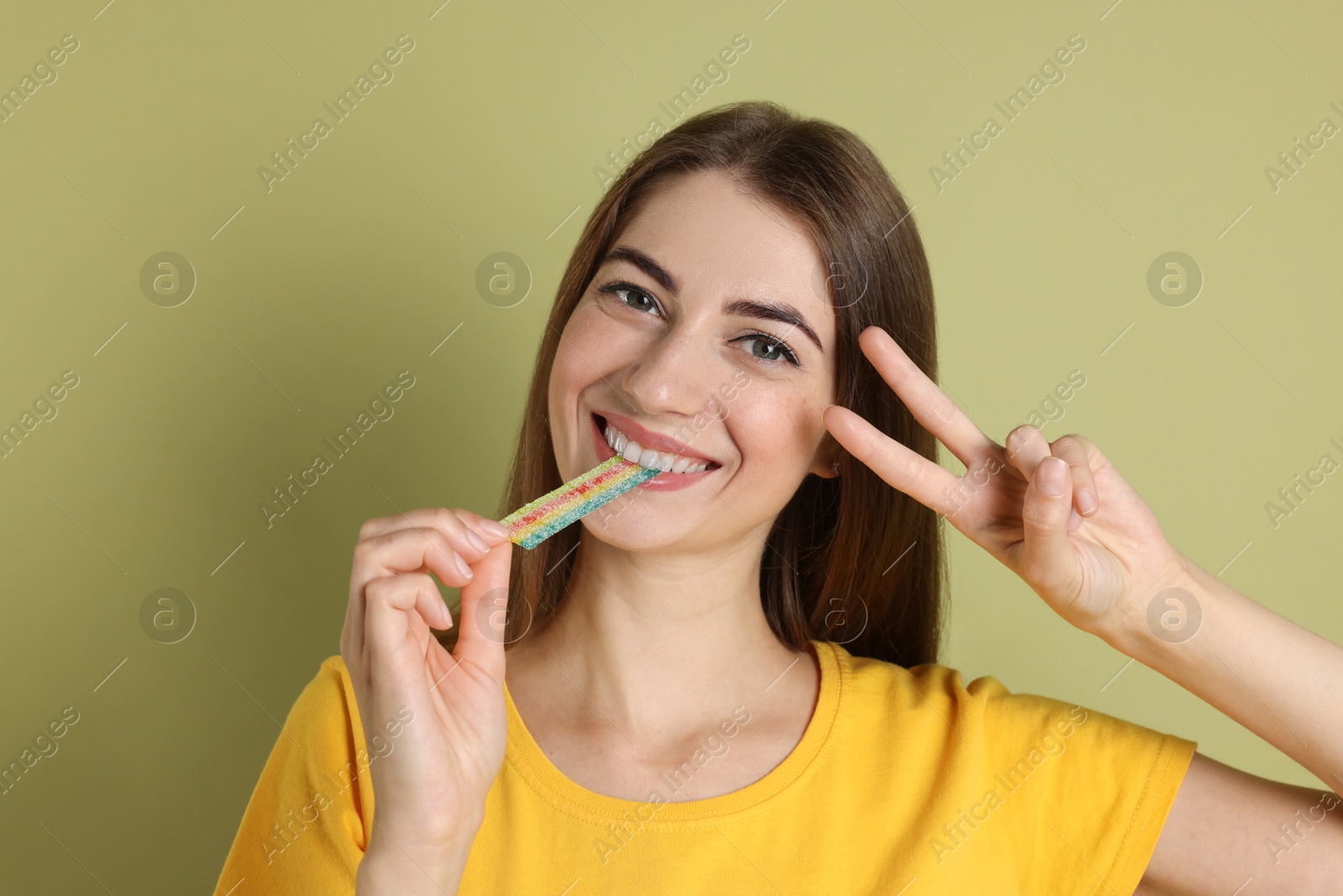 Photo of Young woman eating tasty rainbow sour belt while showing v-sign on olive background